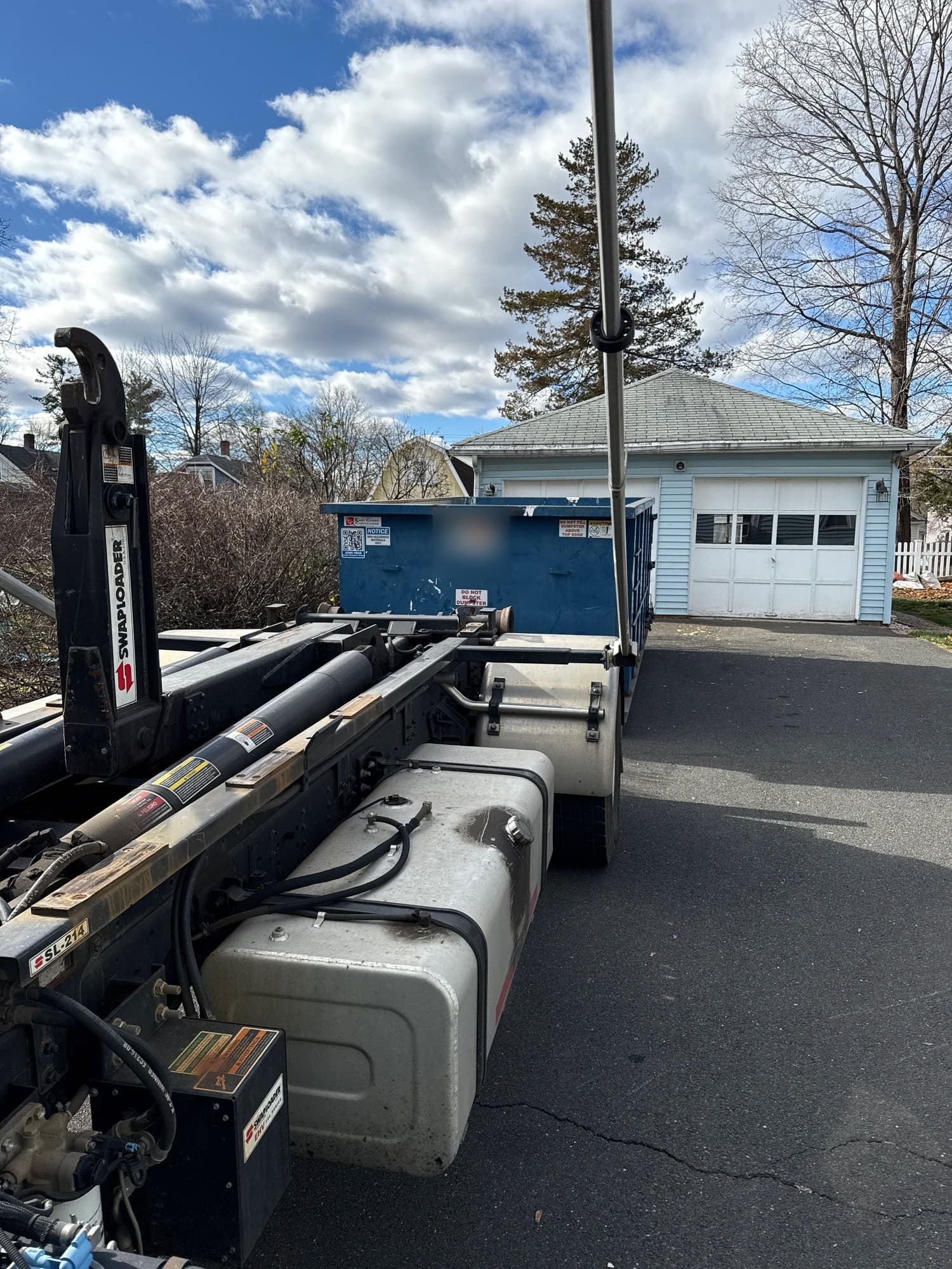 Hook-lift truck and a blue roll-off dumpster in front of a detached garage