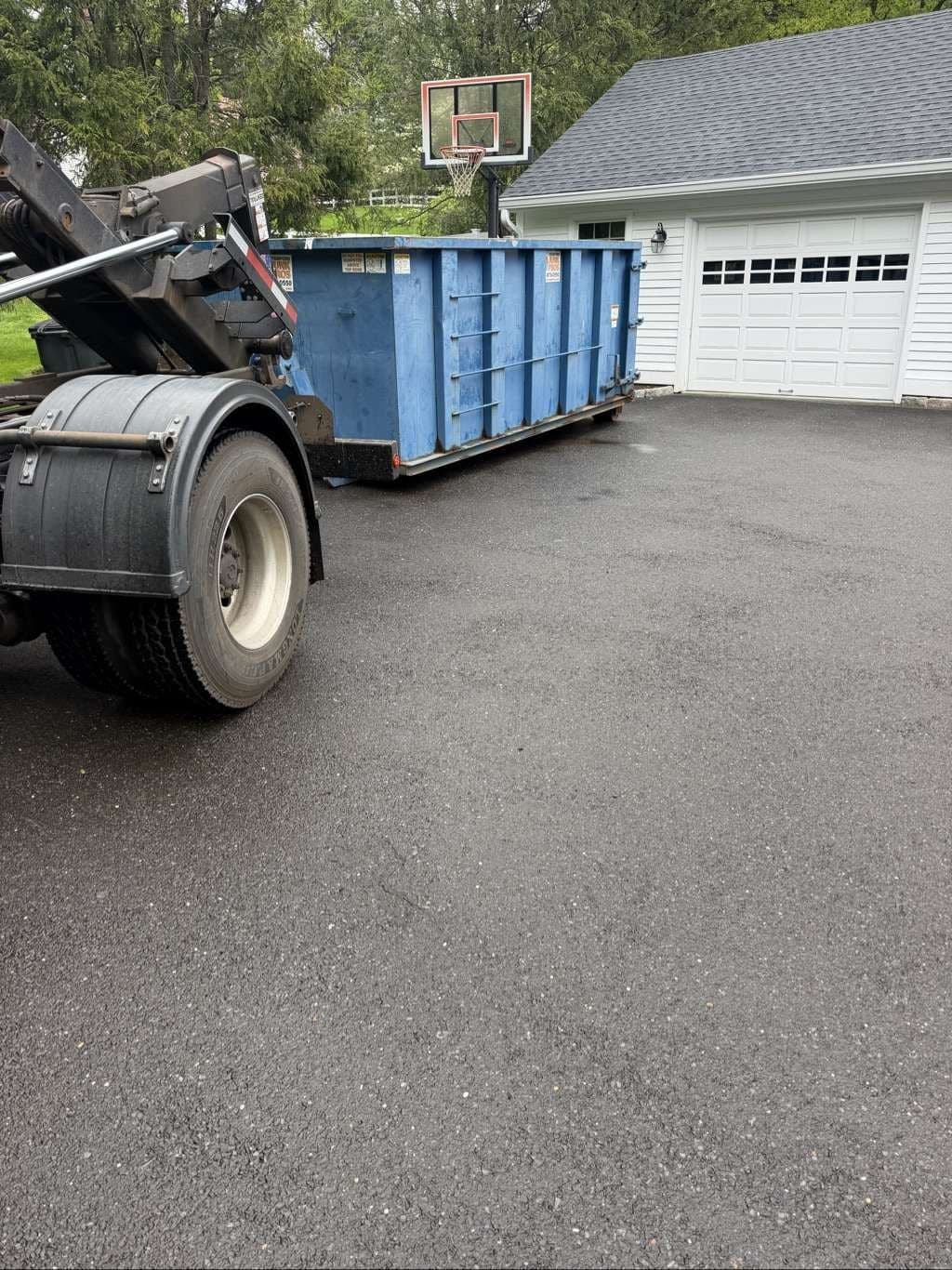 Roll-off truck placing a blue dumpster on a driveway by a garage