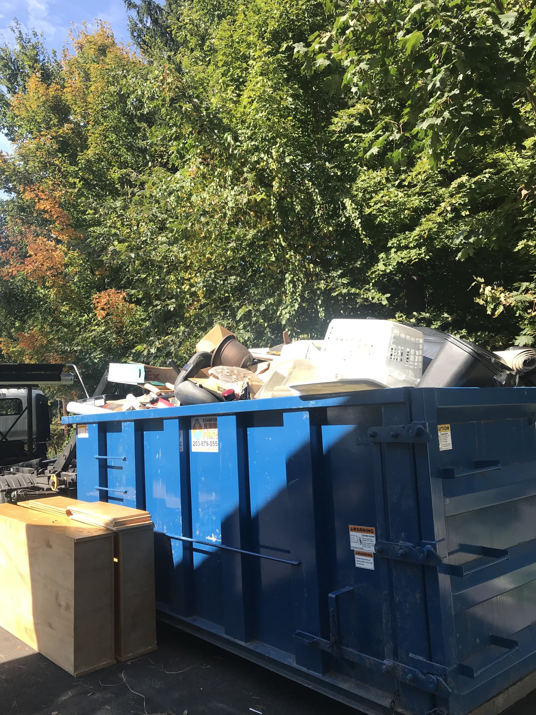 blue roll off dumpster filled with mixed junk and debris trees in background by Grizzly Junk Pros